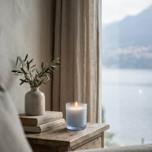 Lake Garda Duftkerze von AVA & MAY auf Holzkommode am Fenster mit Seeblick – frischer Raumduft aus Grünem Tee, Gurke und Maiglöckchen.